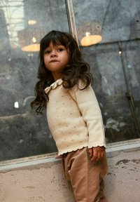 Maglione lavorato a maglia beige chiaro con scollatura a onde e motivo a occhiello, abbinato a pantaloni jogger marrone chiaro, in piedi accanto a una finestra.