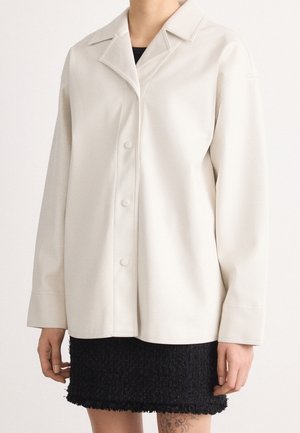 Person iført en off-white knapjakke med lange ærmer over et sort struktureret skørt, der viser en tatovering på venstre lår.