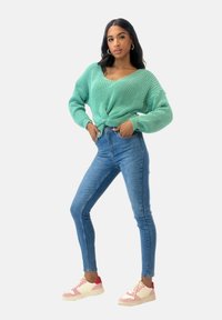 Mintgrüner Strickpullover mit V-Ausschnitt und verdrehtem Vorderteil, kombiniert mit eng anliegenden blauen Jeans und zweifarbigen Sneakern.