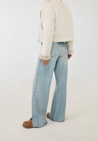 Světlá béžová semišová krátká bunda s kožíškovou podšívkou a manžetami, doplněná o světle modré široké džíny s vysokým pasem a hnědé nazouvací boty.