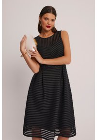 Czarna sukienka w paski z dopasowanym gorsetem i rozkloszowanym dołem, z teksturowanej tkaniny i okrągłym dekoltem. Modelka trzyma białą kopertówkę.