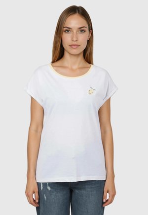 Frau mit glatten braunen Haaren, die ein weißes T-Shirt mit hochgekrempelten Ärmeln und einem kleinen Fruchtmotiv auf der linken Brust trägt, kombiniert mit blauen Jeans.