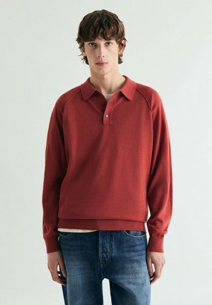 Joven con suéter polo de manga larga color rojo óxido y jeans azules, de pie contra un fondo claro y sencillo.