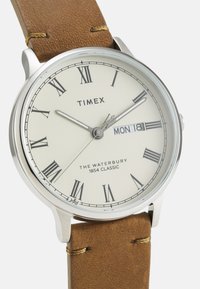 Reloj de pulsera de acero inoxidable con esfera color crema, números romanos, agujas plateadas y correa de cuero marrón. La pantalla de fecha muestra el día y la fecha.
