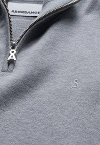 Grau gestrickter Pullover mit Reißverschluss am Kragen und silberner Hardware. Verfügt über ein dezentes, in passendem Grau gesticktes Logo auf der linken Seite.