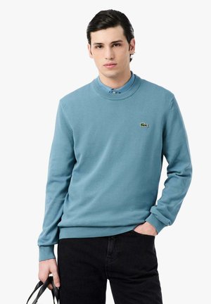Jeune homme portant un pull bleu par-dessus une chemise bleue à col, pantalon noir, avec une main dans la poche et tenant les bretelles d’un sac noir de l’autre.