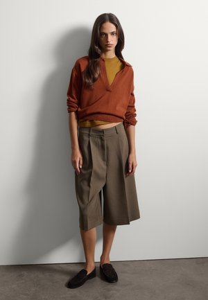 Junge Frau mit langen braunen Haaren trägt einen rostfarbenen Pullover über einem senffarbenen Top, olivgrüne weite knielange Shorts und schwarze Loafers, steht vor einer weißen Wand.