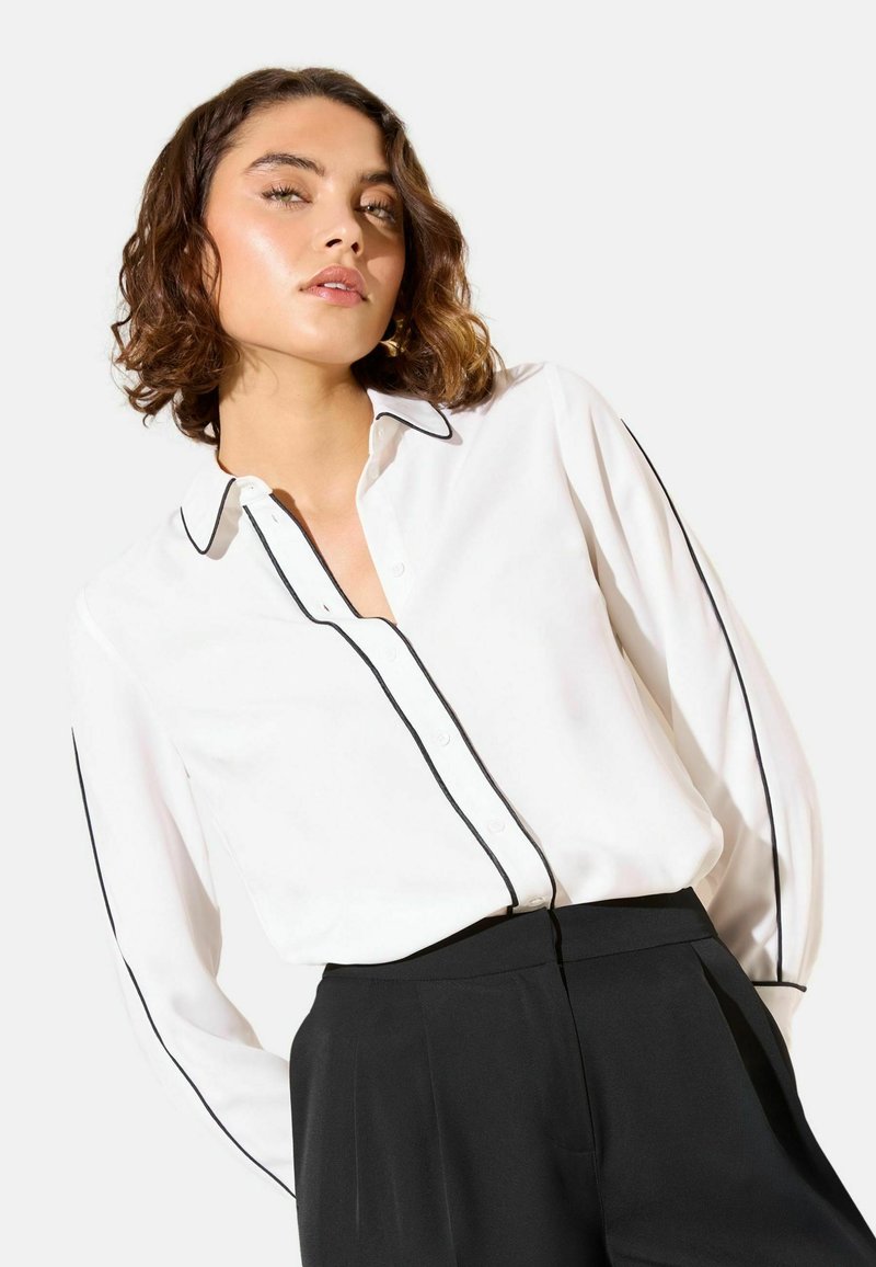 Biela košeľa na gombíky s čiernym lemovaním pozdĺž goliera a rukávov, s voľným strihom a ľahkým materiálom. Kombinovaná s čiernymi nohavicami.
