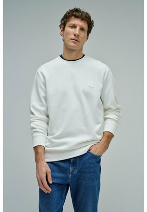 Joven con cabello rizado que lleva una sudadera blanca y jeans azules, posando frente a un fondo gris liso.