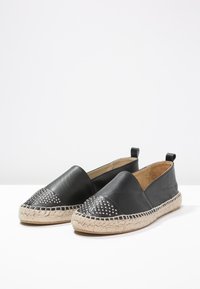 Espadrilles de cuero negras con puntera adornada con tachuelas y suela de yute trenzado. Diseño sin cordones con tirador en la parte posterior. Textura suave con detalles de acento.
