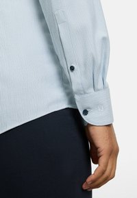 Light blue patterned shirt with a buttoned cuff, featuring dark blue buttons. Smooth texture and tailored design with logo detail on sleeve.