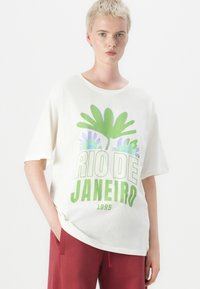 T-shirt surdimensionné de couleur crème avec un graphique floral vert et violet et un texte en gras indiquant "RIO DE JANEIRO 1995". Texture en coton lisse.