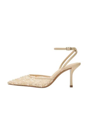 Bézs hegyes orrú magassarkú, virágmintás csipkével borítva, vékony bokapánttal és csatos záródással, fehér háttéren.