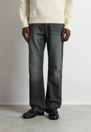 Homme portant un jean noir délavé droit, des mocassins en cuir noir et un pull en maille crème, debout sur un sol gris.