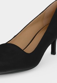 Escarpin noir à talon moyen avec un bout arrondi, une texture lisse et une doublure intérieure beige visible sur fond blanc.