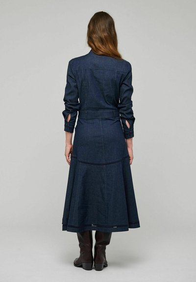 Femme aux longs cheveux bruns, portant une robe en denim bleu foncé à manches longues et des bottes marron montantes jusqu'aux genoux, debout de dos sur un fond uni.