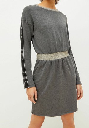 Vrouw draagt een grijze jurk met lange mouwen, een elastische taille en een zwarte streep met witte tekst die over de mouwen loopt.