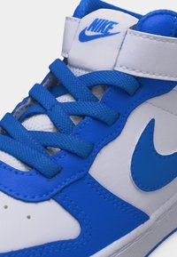 Λευκό και μπλε αθλητικό παπούτσι Nike με μπλε κορδόνια, το σήμα Nike στο πλάι και λογότυπο στη λευκή λουρίδα της γλώσσας.