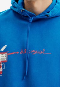 Modrá mikina s kapucí z měkké látky s nastavitelnou šňůrkou, na přední straně červené logo "desigual" a grafické štítky.