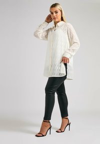 Camicia bianca a maniche lunghe con bottoni e motivo testurizzato, abbinata a pantaloni neri aderenti e tacchi a strappo neri, in piedi davanti a uno sfondo neutro.