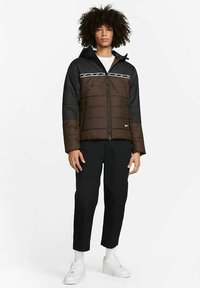 Giacca puffer marrone e nera con cappuccio, caratterizzata da un logo bianco e dettagli a righe, indossata con pantaloni neri e sneakers bianche.