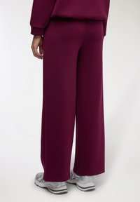 Pantaloni larghi in tessuto di un profondo bordeaux, caratterizzati da una texture liscia e una cintura elastica. Abbinati a scarpe da ginnastica grigie.