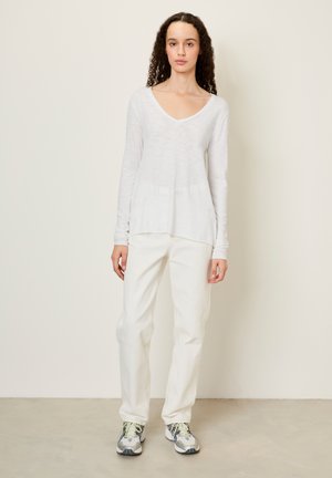 Hvid langærmet V-hals top lavet af letvægtsstof, parret med hvide straight-leg jeans og grå sportsko med neonlysninger.