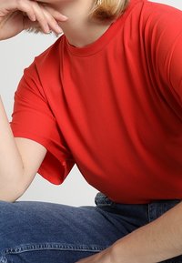 T-shirt rossa a maniche corte con collo rotondo e texture liscia, abbinata a jeans blu. Il tessuto sembra leggero e informale.