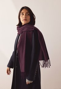 Vīna sarkana apļveida šalle no mīksta auduma, pārklāta pār tumšu mēteļa. Ir aprīkota ar bārkstīm un slāņotu dizainu, kas uzlabo siltumu un tekstūru.