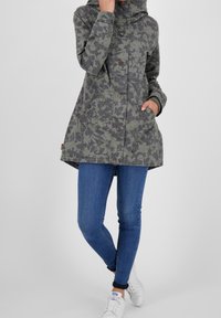 Chaqueta verde con estampado floral, con capucha, cierre de botones y dos bolsillos delanteros, llevada sobre jeans azules y zapatillas blancas.