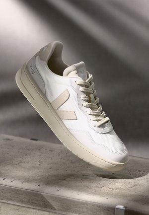 Sneaker bianca e beige con lacci e marchio "V-90", mostrata fluttuare sopra una superficie di pietra grigia con sfondo ad ombra soffusa.