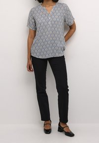 Blå kortærmet top med et dekorativt creme- og guldmønster, kombineret med sorte tætsiddende bukser og sorte sko med blokhæl.