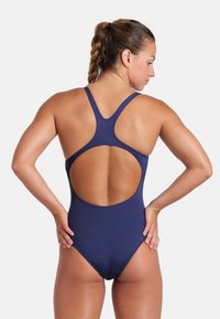 Femme aux cheveux tressés portant un maillot de bain une pièce bleu marine avec un dos nu et des bretelles croisées, se tenant les mains sur les hanches, tournée de profil.