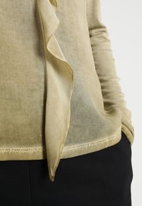 Blusa verde oliva chiaro con una texture trasparente, dettaglio arricciato sul davanti e orlo cucito. Indossata sopra un indumento nero.
