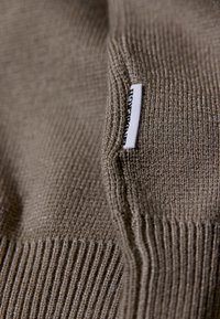 Kudottu ruskea kangas, jossa on uurteinen rakenne ja pieni valkoinen lappu mustalla tekstillä, sijoitettuna sauman lähelle.