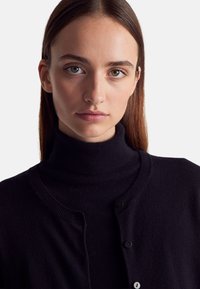 Černý rolák vyrobený z měkkého materiálu, s ribbovým límcem, zapínáním na knoflíky a jemnou texturou, která zdůrazňuje elegantní, přiléhavý střih.