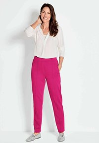 Fuchsia broek met zijzakken en voornaaddetails, gecombineerd met een witte blouse. Vrouw staat rechtop met een glimlach en draagt zilveren schoenen.