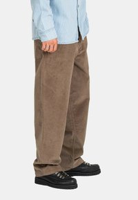 Persona vestida con pantalones anchos de corduroy marrón, zapatos negros con cordones y una camisa de manga larga a rayas azul y blanca, de pie contra un fondo blanco.