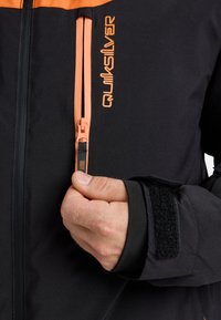 Giacca nera con accenti arancioni, dotata di cerniera frontale e chiusura con polsino in Velcro. Il tessuto appare liscio e resistente. Logo Quiksilver visibile.