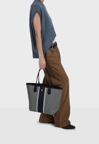 Černá a vzorovaná taška typu tote s dvojitými uchy, s texturovanými boky a centrálním černým proužkem. Nenošeno s neformálním šedým topem a hnědými kalhotami.