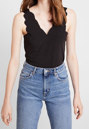 Femme portant un haut noir sans manches avec un col festonné et un jean bleu taille haute, debout devant un fond uni.