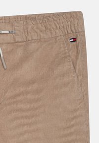 Beige corduroybroek met elastische tailleband, koord met merkembleem, zijzak en klein rechthoekig logo in rood, wit en blauw.