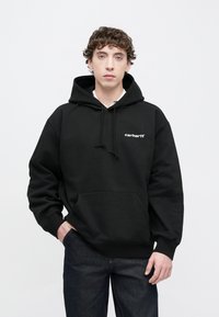 Sudadera negra con capucha, bolsillo tipo canguro y cordones. Presenta un logo blanco en el pecho. Combinada con pantalones vaqueros oscuros.