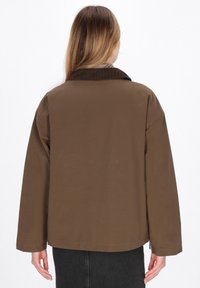 Brun, oversized jakke med en kordfløjlskrave, lange ærmer og en glat tekstur. Har en lynlåsdetaile ved kanten.