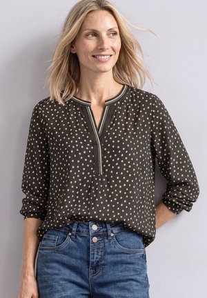 Femme aux cheveux blonds portant un chemisier noir à manches longues à pois et un jean bleu, souriante et regardant sur le côté sur un fond uni.