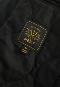 Svart quiltad tyg med en guld- och svart etikett som skriver "NEXT", pryds av ett emblem, storlek "M" och texten "Tillverkad i Kina".