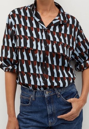 Personne portant une chemise ample à boutons avec un motif géométrique noir, bleu clair et marron, rentrée dans un jean bleu taille haute.