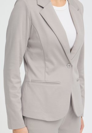 Femme portant un blazer gris clair ajusté par-dessus une chemise blanche, montrant les détails des boutons et des poches à l’avant, cadrée au niveau du cou et des hanches.