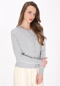 Cardigan lavorato a maglia grigio chiaro con scollatura rotonda, dotato di bottoni dorati e maniche lunghe. Texture morbida e design aderente.