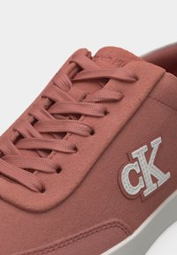 Gros plan sur une basket rose poussiére avec des lacets assortis, une semelle en caoutchouc blanche et un logo "cK" blanc en relief sur le côté.